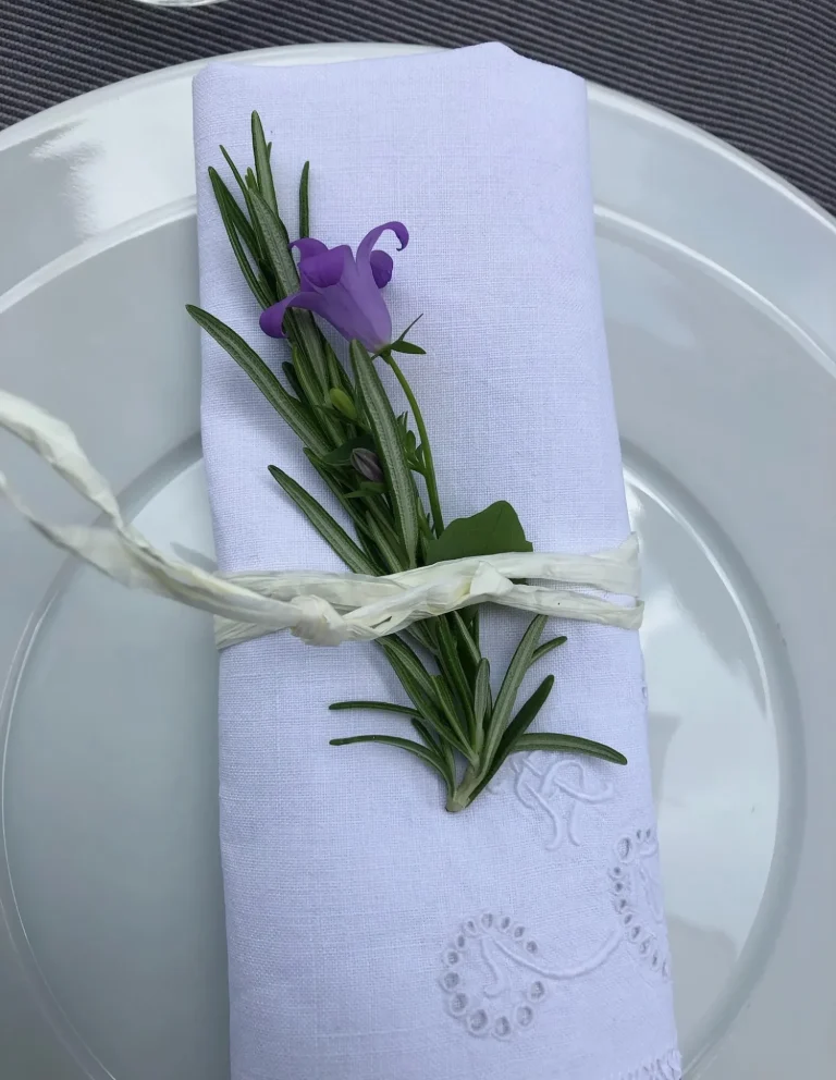Stoffserviette mit Rosmarin und Blüte – stilvolle Tischdekoration Weiße Stoffserviette mit Rosmarinzweig und violetter Blüte, dekorativ auf einem weißen Teller angerichtet.